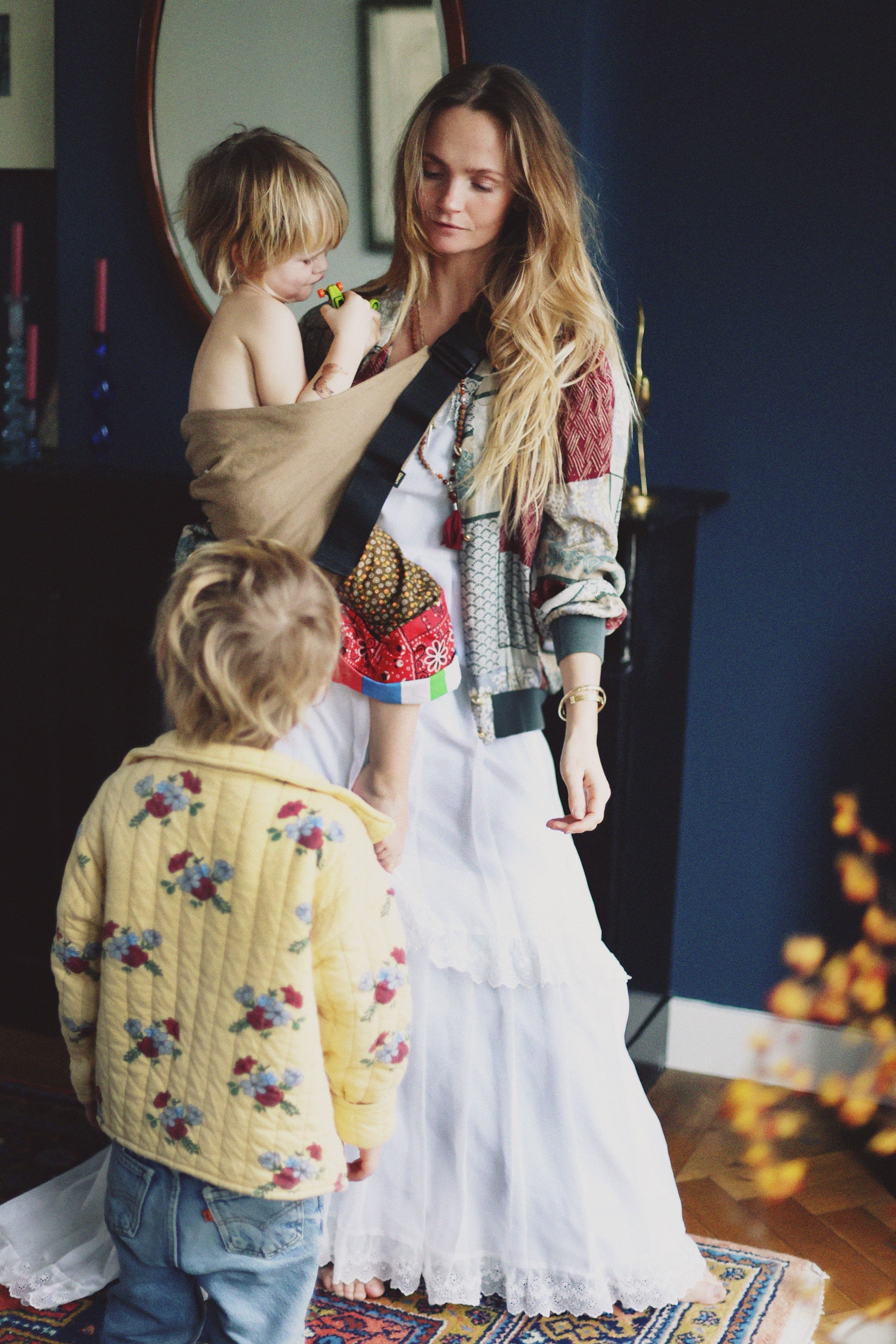 A mother wearing the Wildride Camel toddler carrier at home, holding a child while another stands nearby.