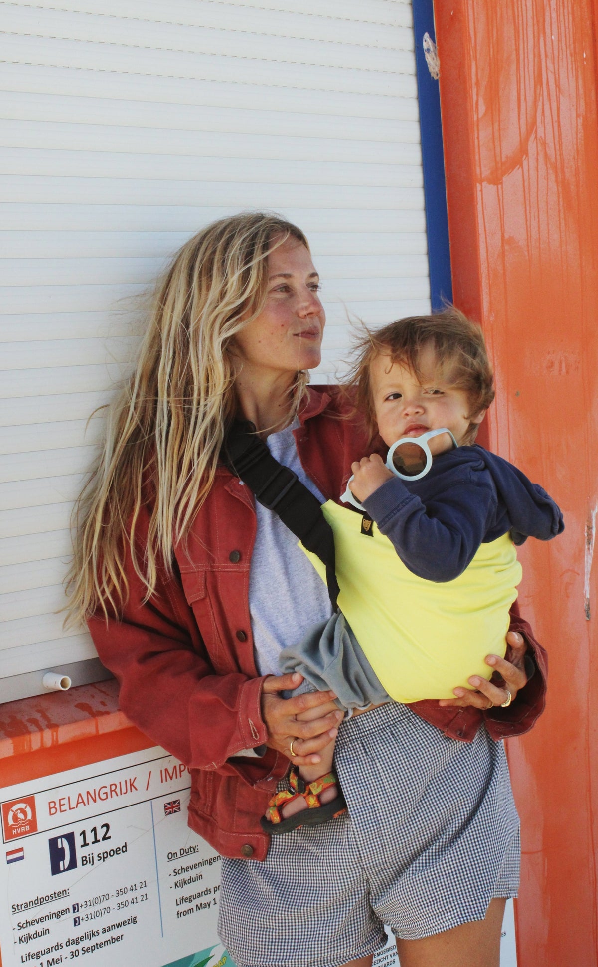 A mother wears a Wildride toddler carrier in Lemon, holding her child close in a hip carrier. The toddler, in a navy hoodie, looks at the camera while gripping a pacifier. The best baby carrier is secured with a black strap, offering hands-free comfort.