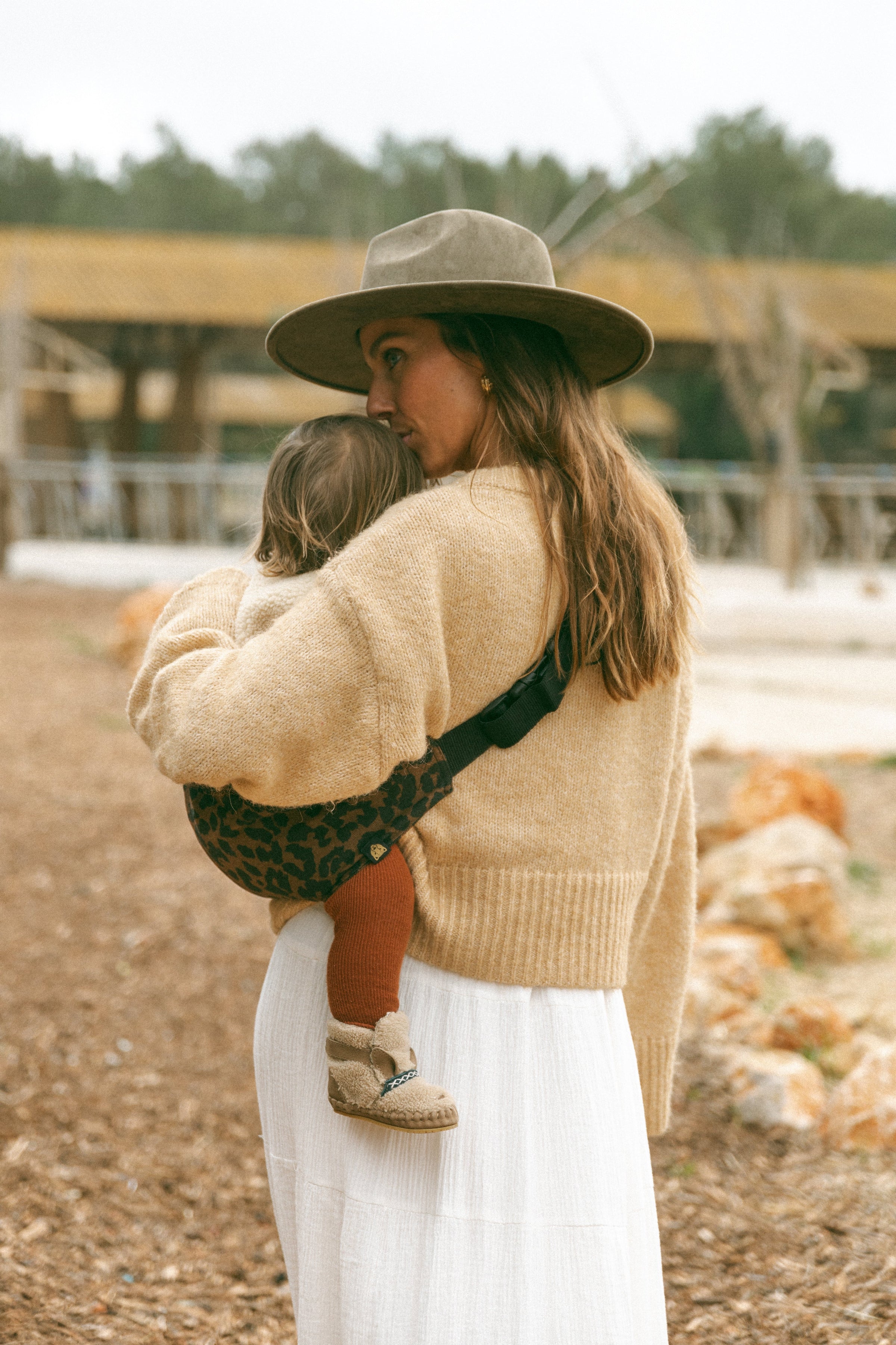 Woman in a beige sweater and hat carrying a toddler on her hip in a Wildride brown leopard print toddler carrier — ergonomic hip carrier for everyday use