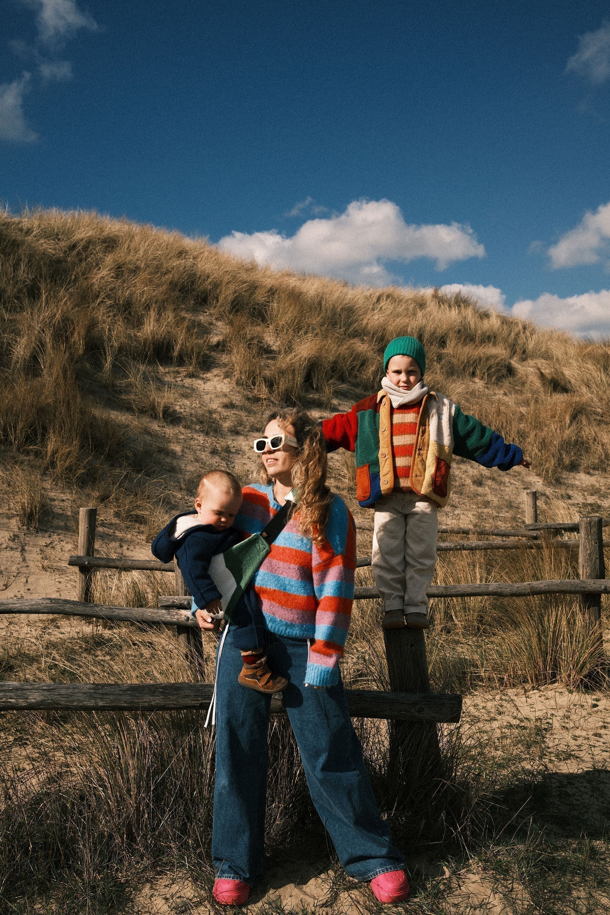 Mother standing in the dunes with her baby in a Wildride baby carrier, while her older child balances on a wooden fence nearby. A calm family moment with their toddler carrier.