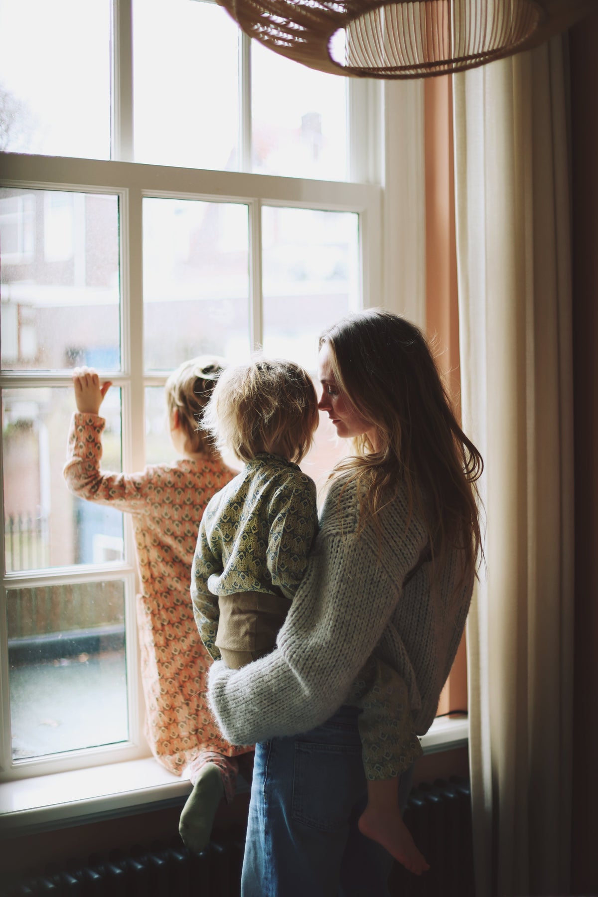 A mother holds her toddler in a Wildride toddler carrier while standing by a window, with another child looking outside. Natural light fills the room.
