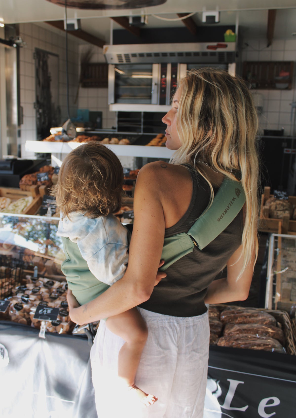 Mother carrying child in Wildride Fern carrier at a bakery counter
